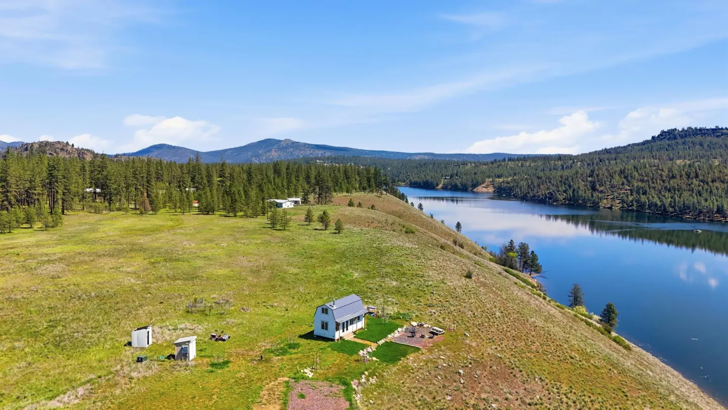 Aerial view of a white house with a grey roof on a grassy hill next to a blue lake and green trees under a blue sky.