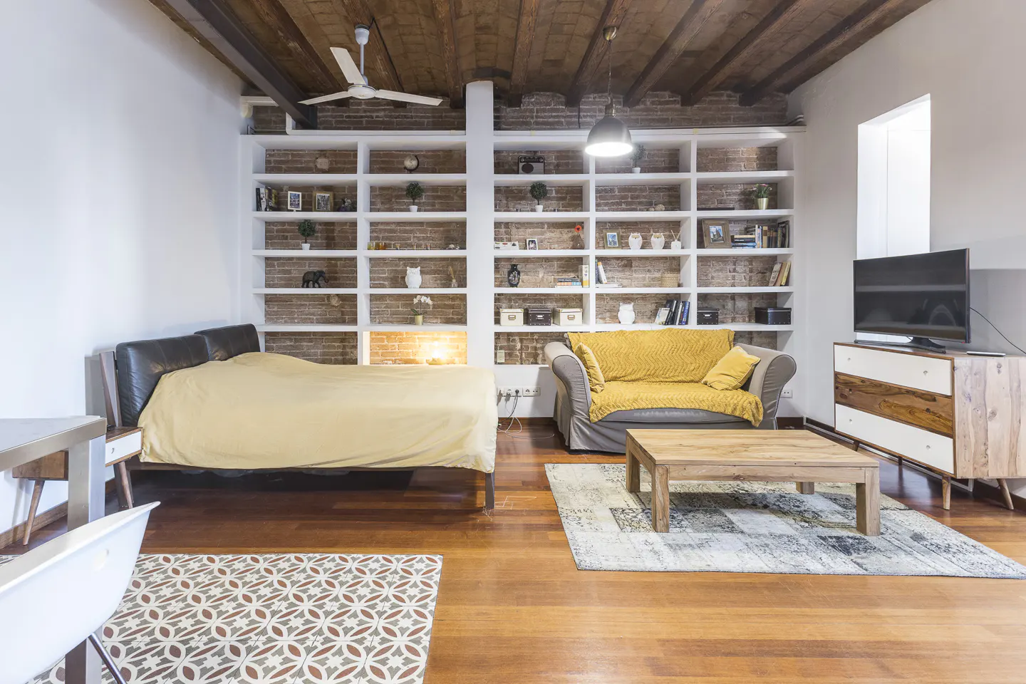 Bright studio apartment with hardwood floors, a bed, a couch, and a large bookshelf filled with decorative items.