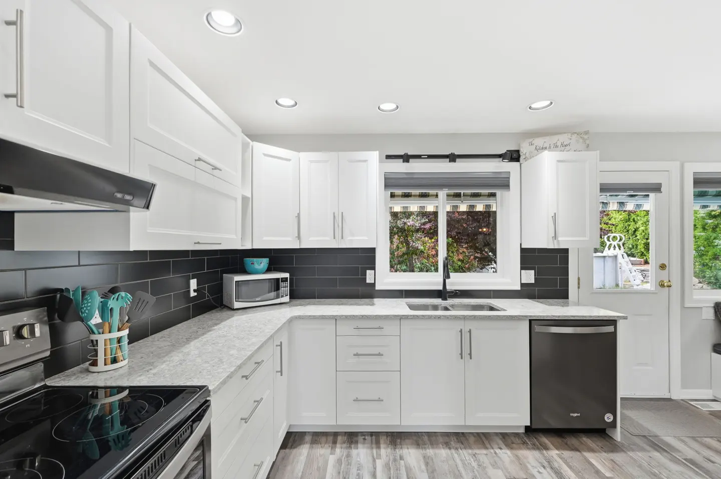 Bright kitchen with white cabinets, black backsplash, and gray countertops. Stainless steel appliances and wood-look flooring. Window view of greenery.