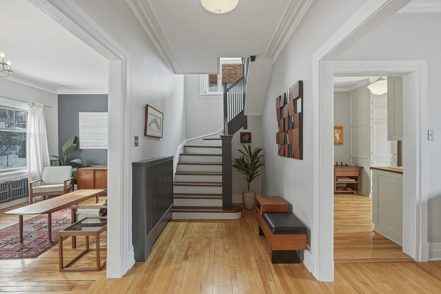 A bright foyer with hardwood floors, a staircase, and doorways to a living room and kitchen. A bench and artwork decorate the space.