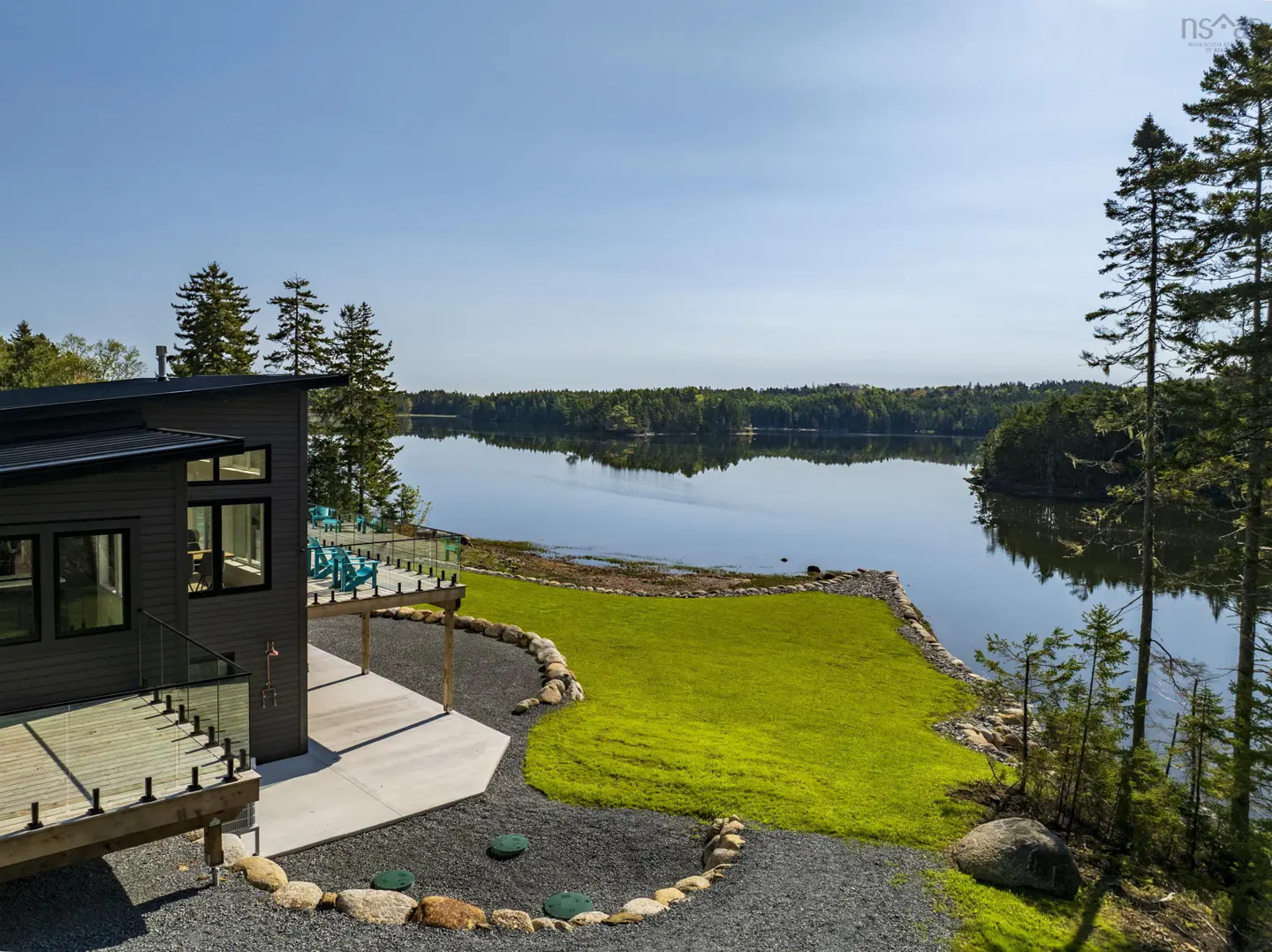 Waterfront home with a gray exterior, wooden deck, and glass railings overlooking a calm lake surrounded by green trees.