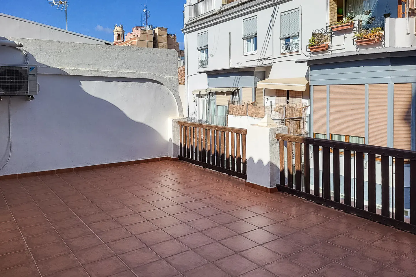 A rooftop terrace with brown tile flooring and a wooden railing, with buildings in the background.