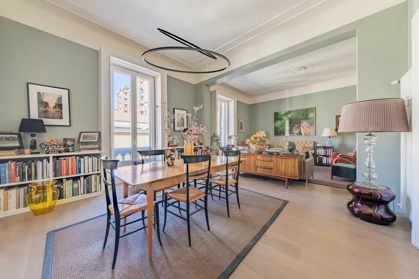 A dining room with light green walls, a wooden table with black chairs, and a modern black light fixture. A bookshelf and a living room are visible in the background.