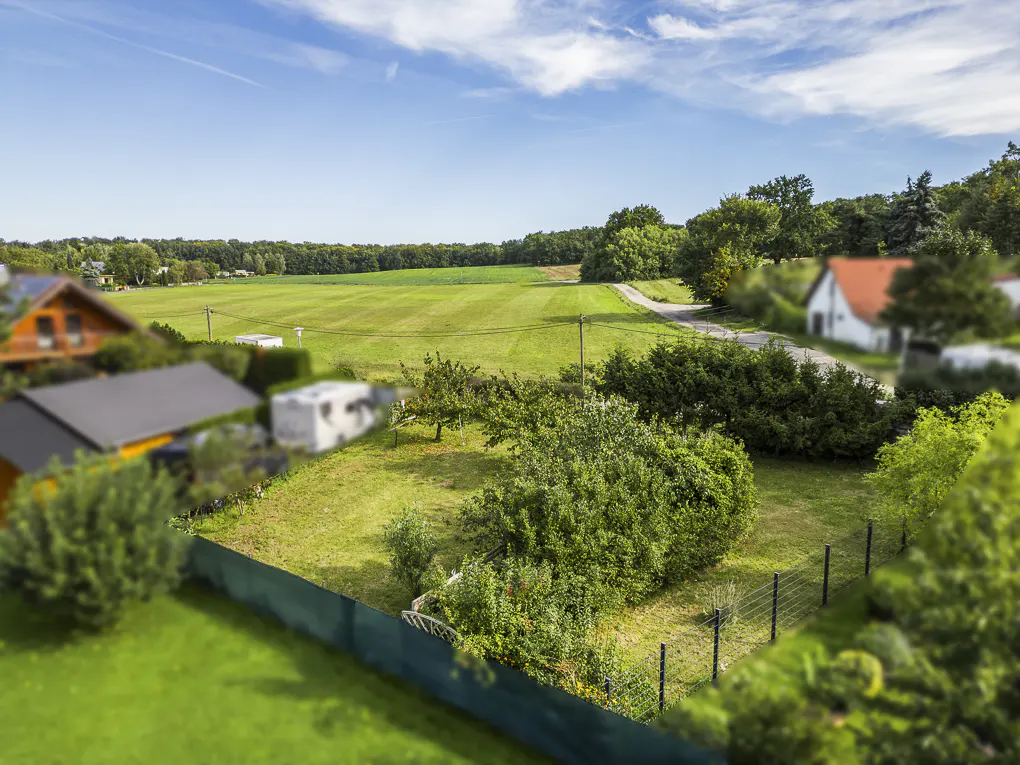 A high angle view of a grassy lot with a fence, trees, and houses under a blue sky.