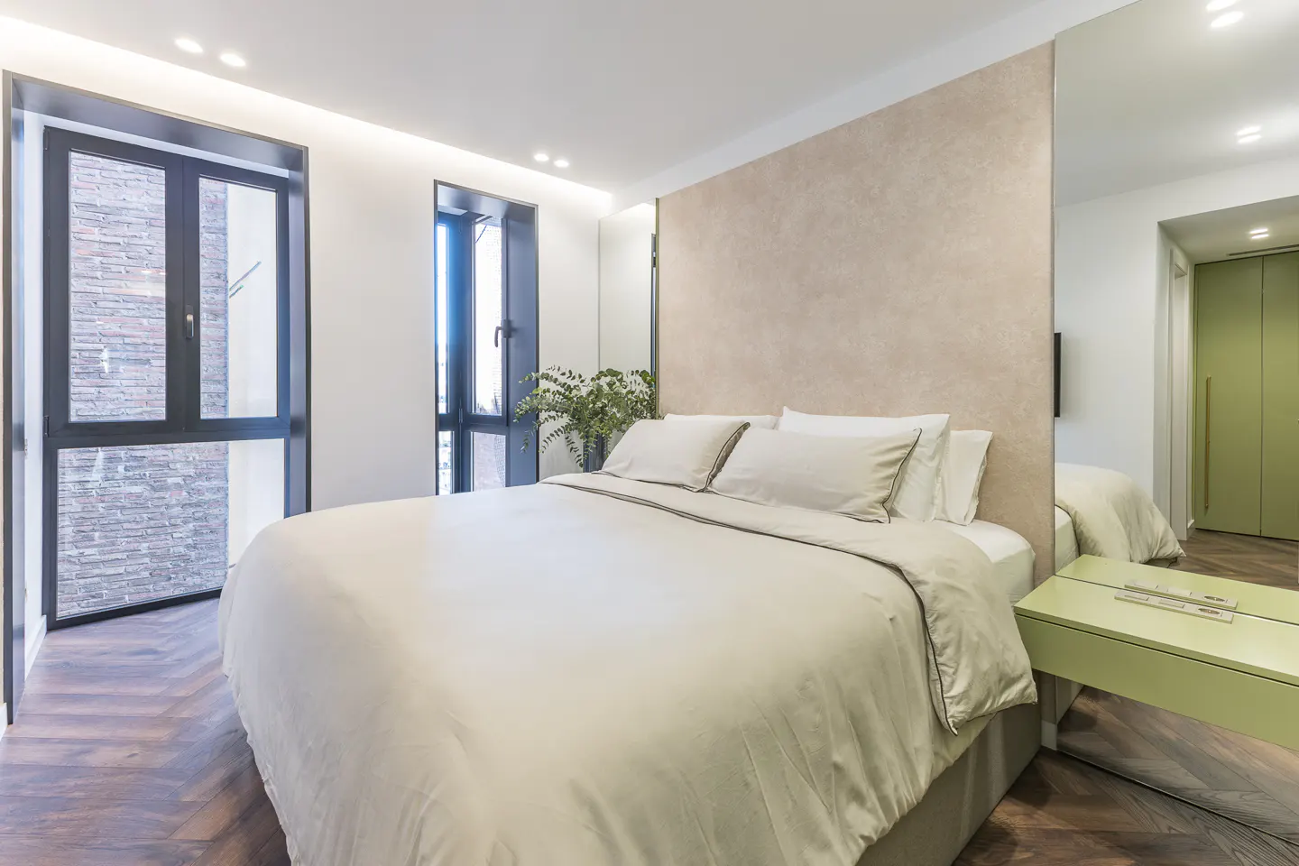 A modern bedroom with a large bed, beige bedding, and a light green nightstand. Two black-framed windows let in natural light. A mirror reflects a green closet.