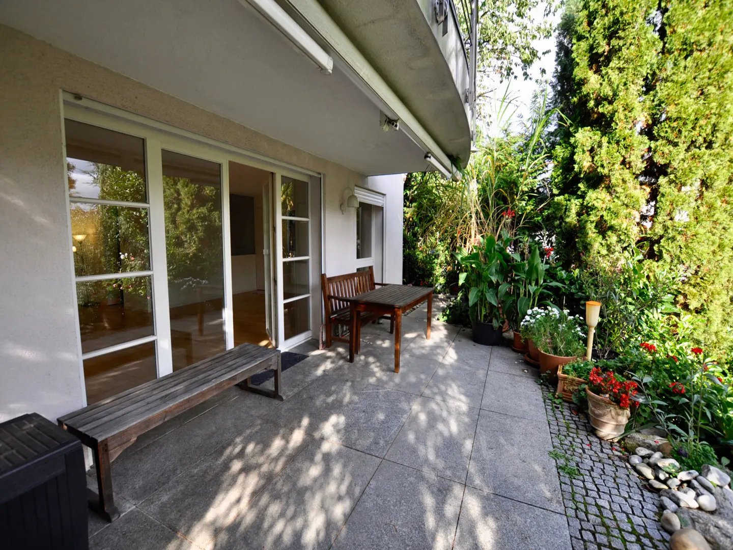 A patio with a wooden bench, table, and chairs sits outside a building with sliding glass doors, surrounded by lush greenery.
