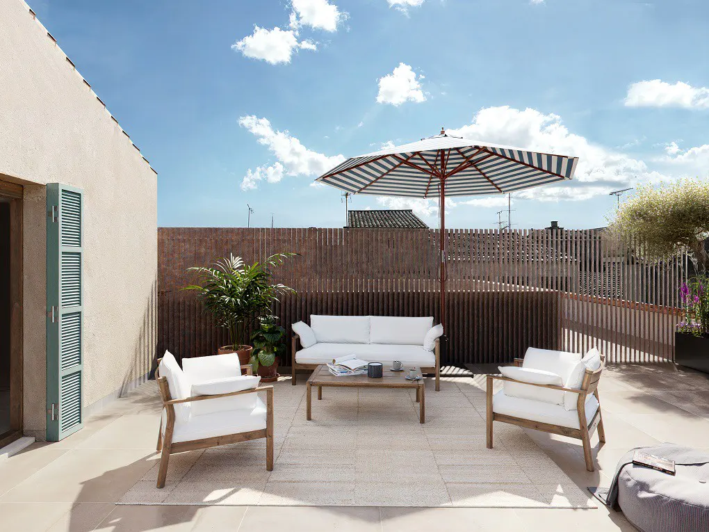 Outdoor patio with white sofa, chairs, and striped umbrella. A wooden fence and blue sky are in the background.