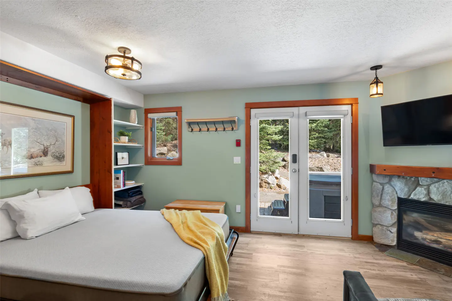 A bedroom with a bed, fireplace, and double doors leading to an outdoor area. The walls are light green, and the floor is wood.