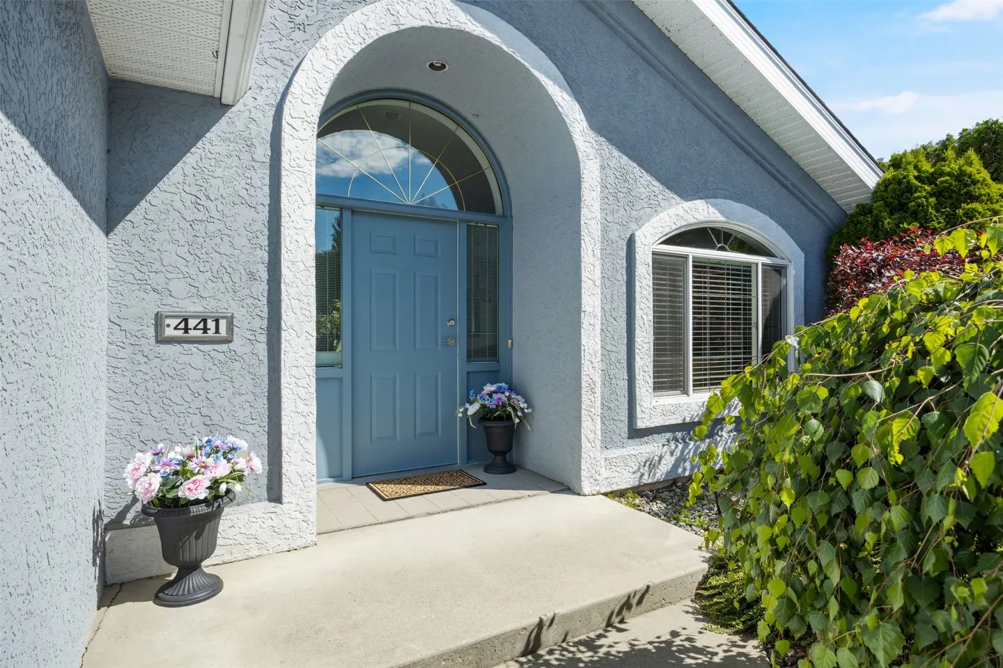 Exterior of a light blue house with a blue front door under an arched entryway. Flower pots flank the door. Address number 441 is visible.