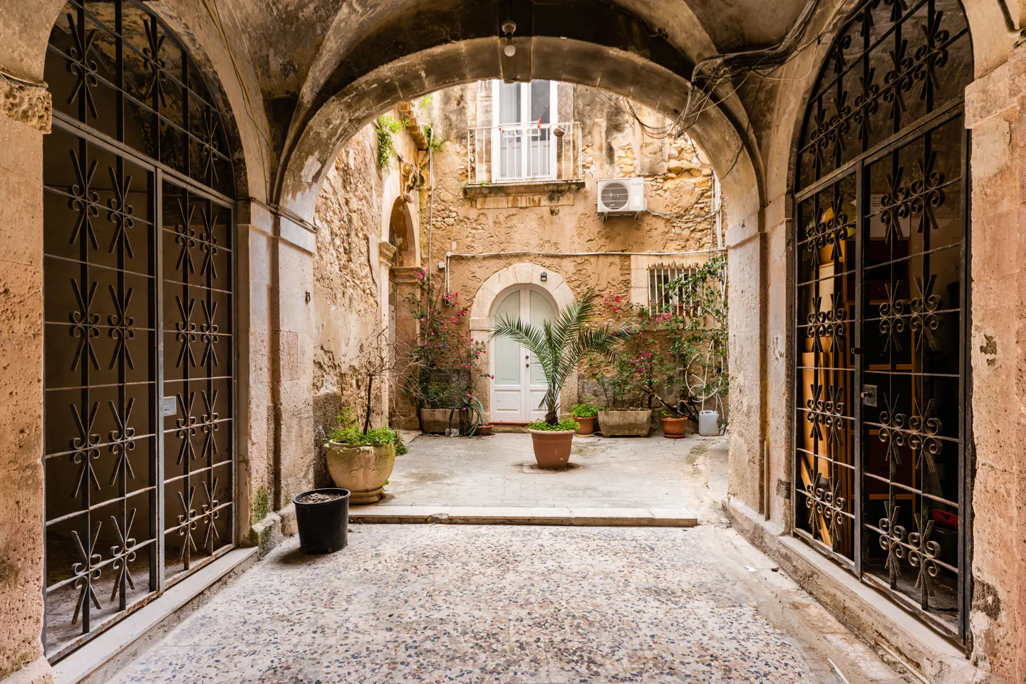 Arched stone entryway to a courtyard with potted plants and a white door. Wrought iron gates flank the entrance.