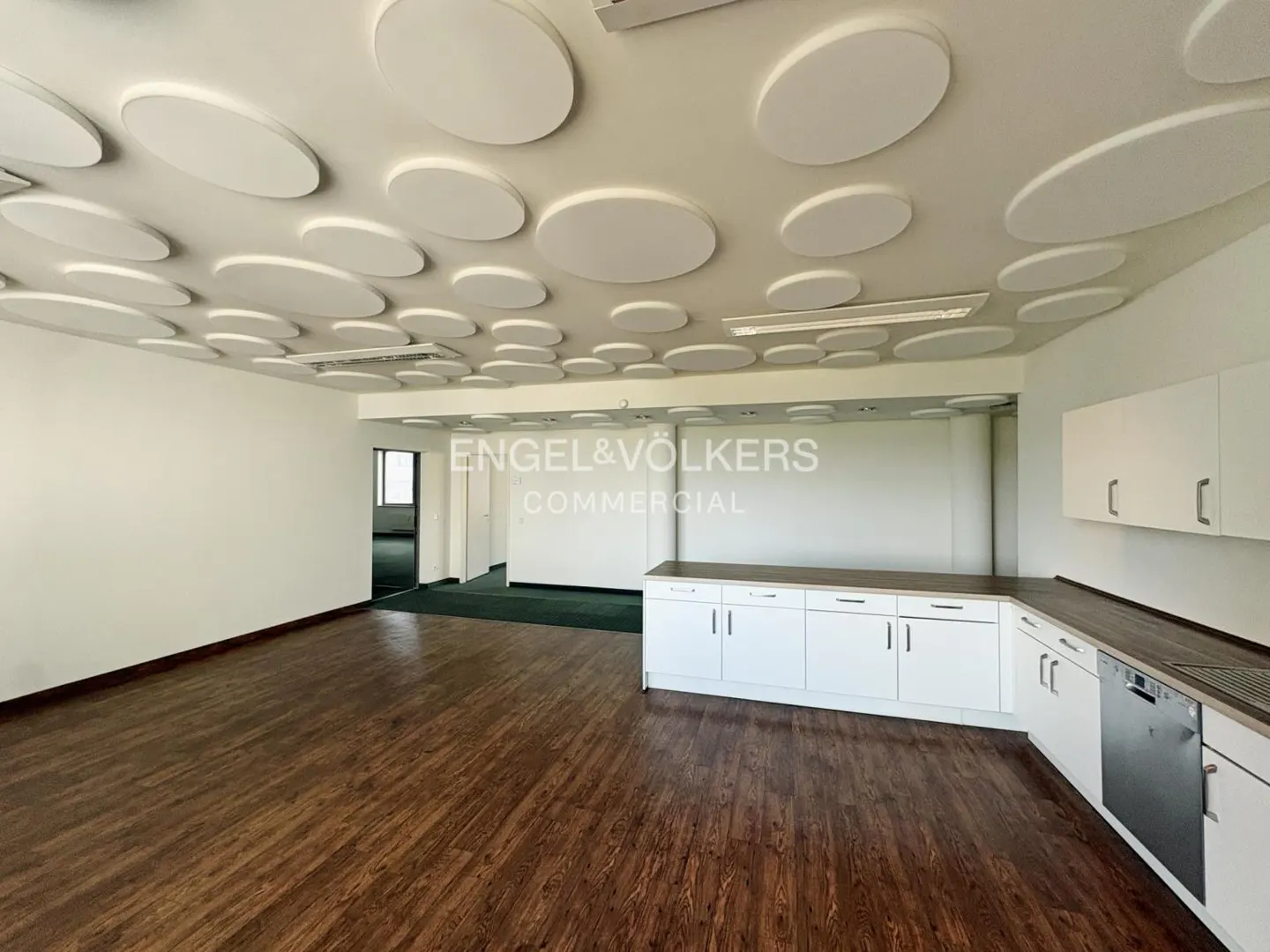 Bright office space with wood floors, white walls, and a ceiling with white circular soundproofing panels. A white kitchen area is in the back.