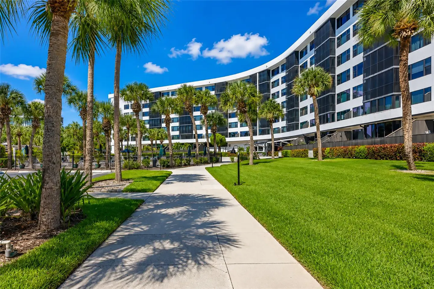Concrete path leading to a modern, curved white apartment building surrounded by palm trees and a lush green lawn.