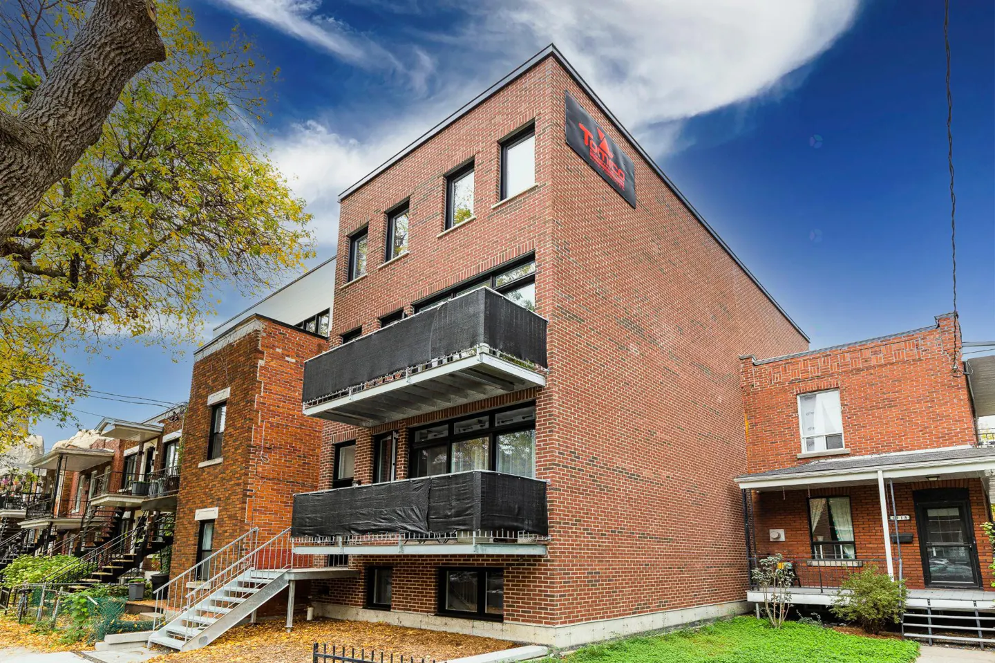 Exterior view of a three-story red brick apartment building with black balconies and windows under a blue sky.
