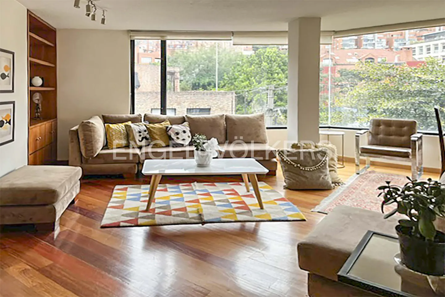 Bright living room with hardwood floors, a large beige sectional, colorful geometric rug, and floor-to-ceiling windows.
