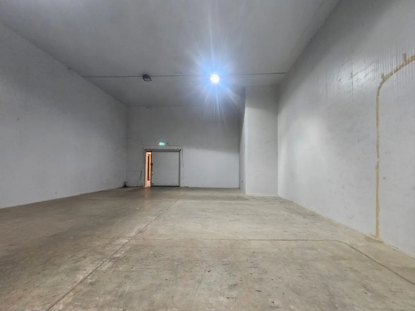 Empty warehouse space with white walls, concrete floor, and a partially open door at the back. A bright light illuminates the room.