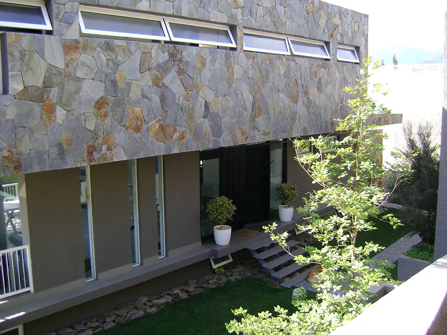 Modern home exterior with a stone facade, gray trim, and a black front door. A small tree and potted plants decorate the entrance.