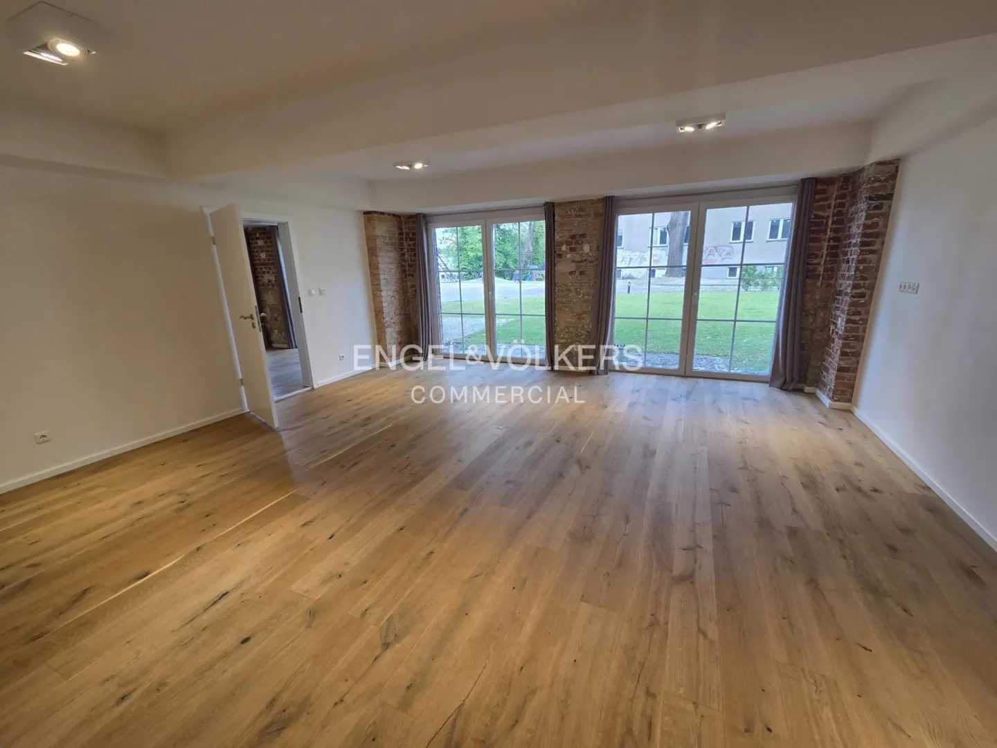 Bright, empty room with hardwood floors, white walls, and brick accents around large windows. Doorway to another room is visible.