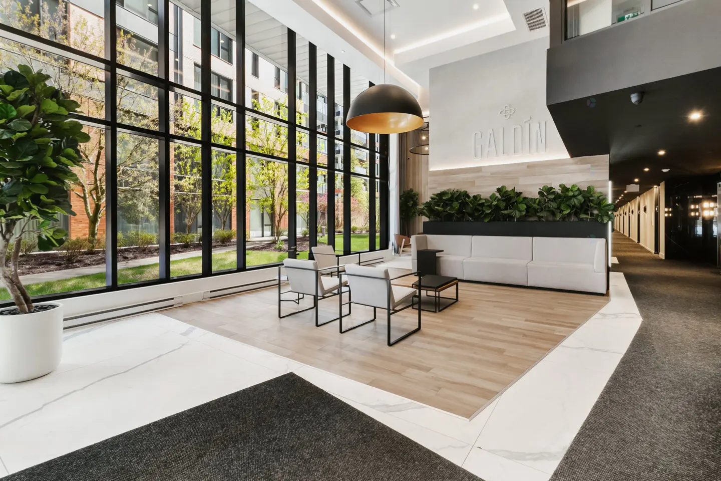 Modern lobby with large windows, chairs, and a white sofa. A black and gold pendant light hangs above. Outside, trees and a building are visible.
