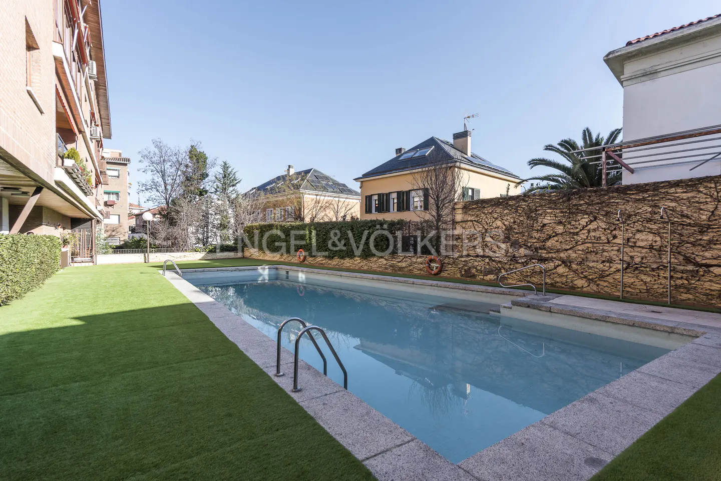 Outdoor pool with blue water, surrounded by green turf and stone. Buildings and a vine-covered wall are in the background.
