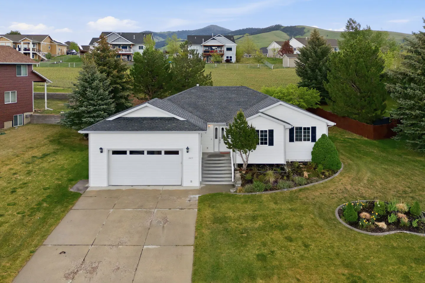 A white, single-story house with a gray roof, a two-car garage, and a concrete driveway. The house is surrounded by a green lawn and trees. Hills are in the background.
