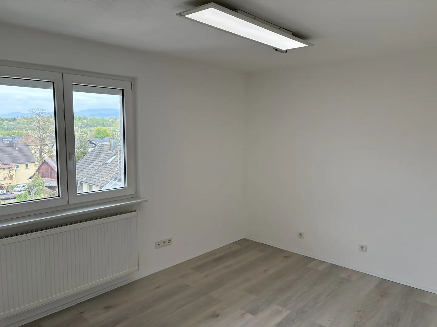 Bright, empty room with white walls, light wood floor, and a window showing a view of houses and trees. A white radiator is under the window.