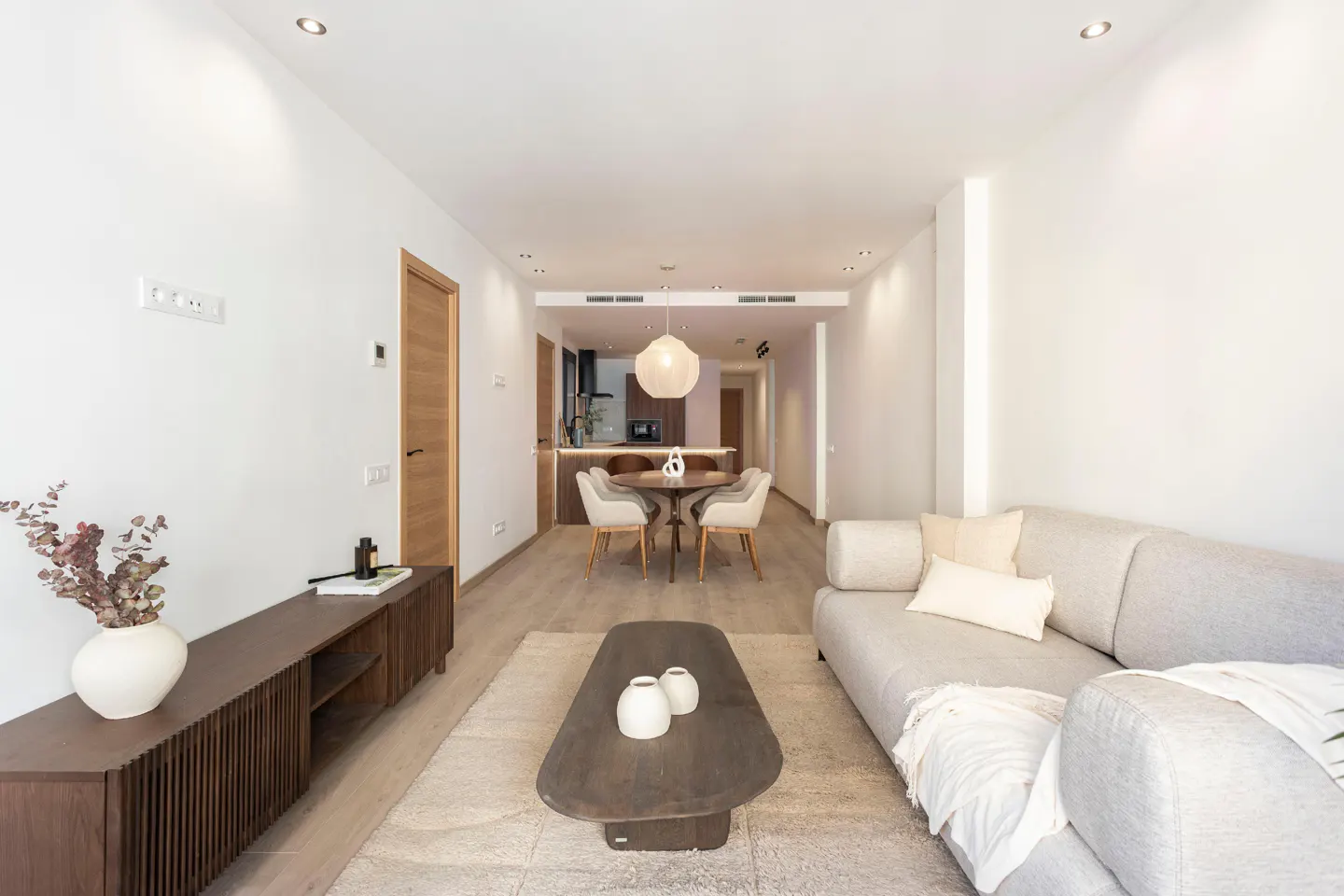Bright, modern living room with a gray sofa, wood coffee table, and TV stand. A round dining table and kitchen are in the background.