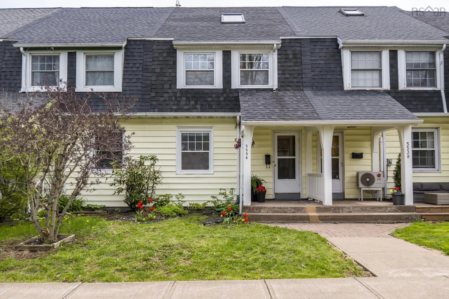 A yellow townhouse with a dark gray roof and white trim. A small lawn and brick walkway lead to the front doors.