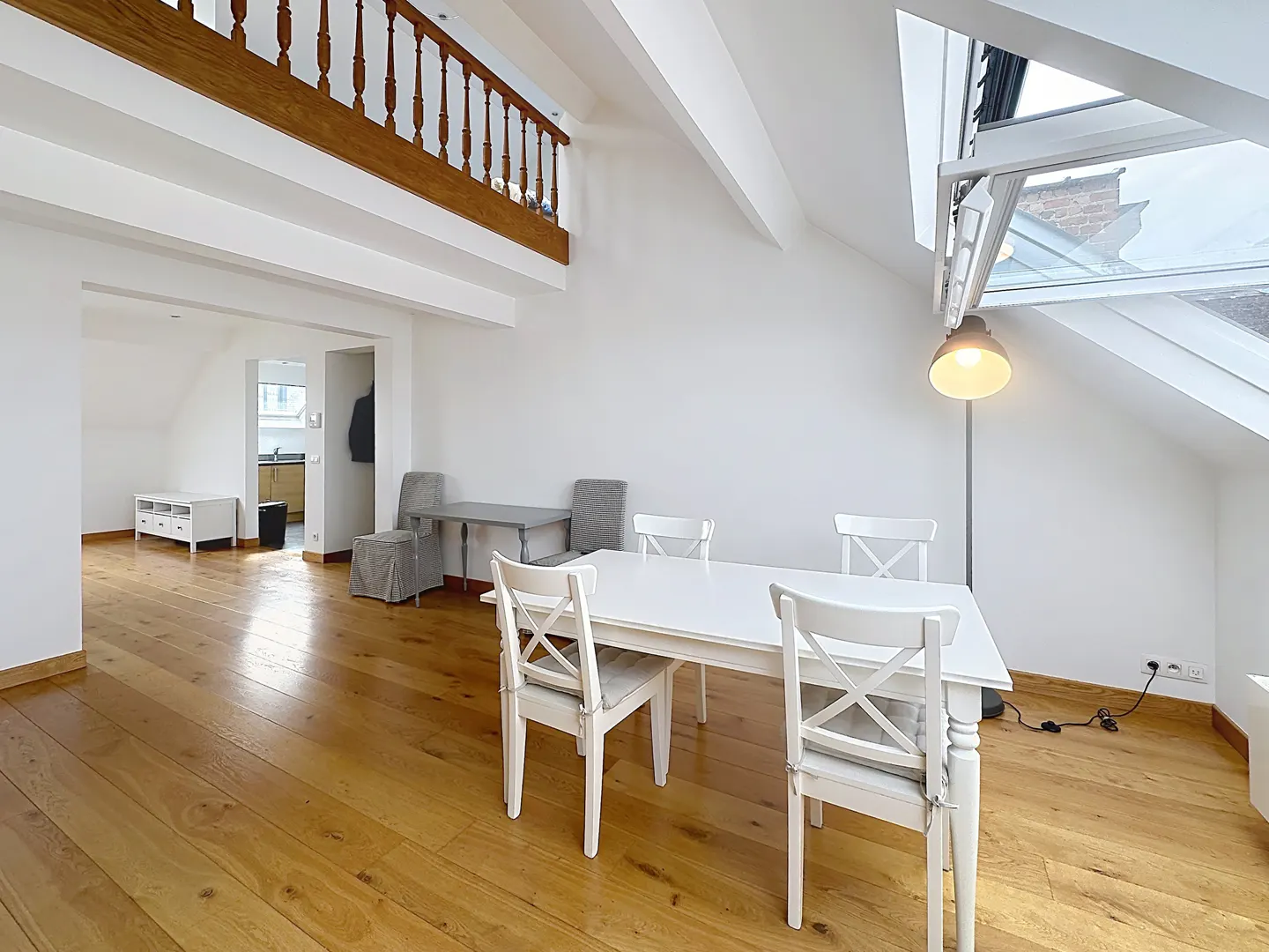 Bright, open-plan apartment with wooden floors, white walls, and a skylight. A white dining table with chairs sits in the center, with a wooden railing above.