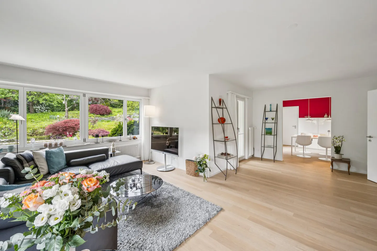 Bright living room with a black leather sofa, glass table, and large windows overlooking a lush garden. A red accent wall is visible in the kitchen.
