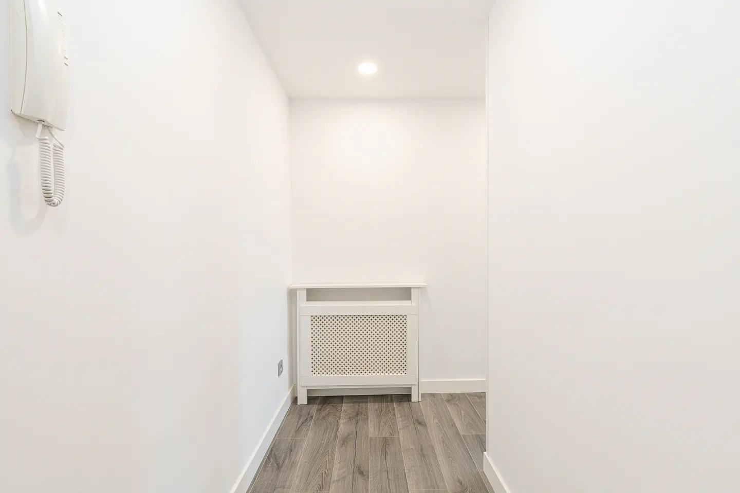 Bright hallway with white walls, gray wood floors, and a white radiator cover. An intercom hangs on the left wall.
