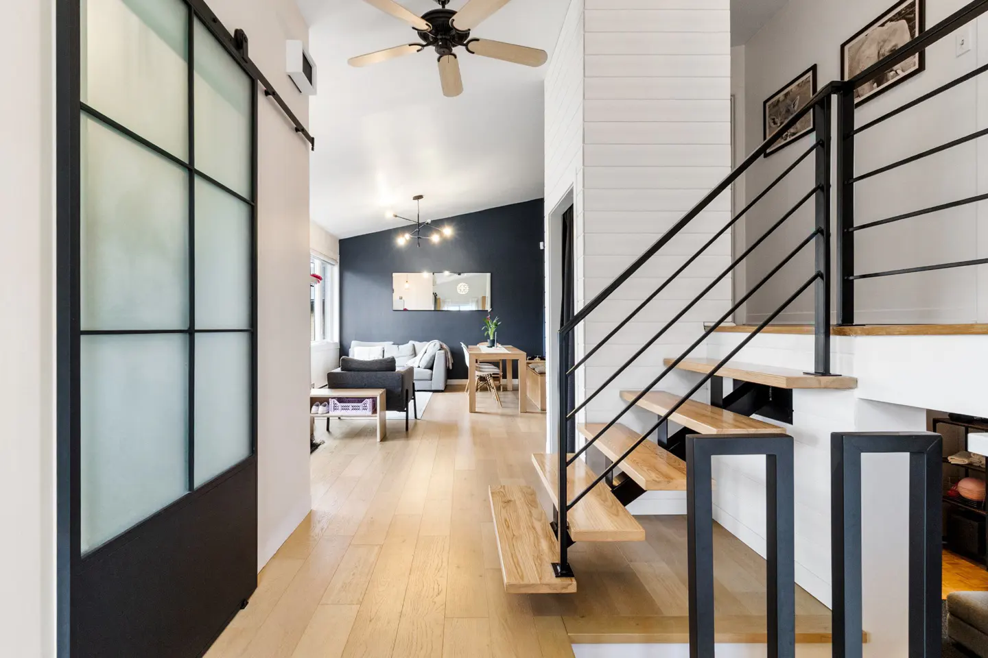 Interior view of a home with light wood floors, stairs with black railings, and a sliding glass door. A living room is visible in the background.