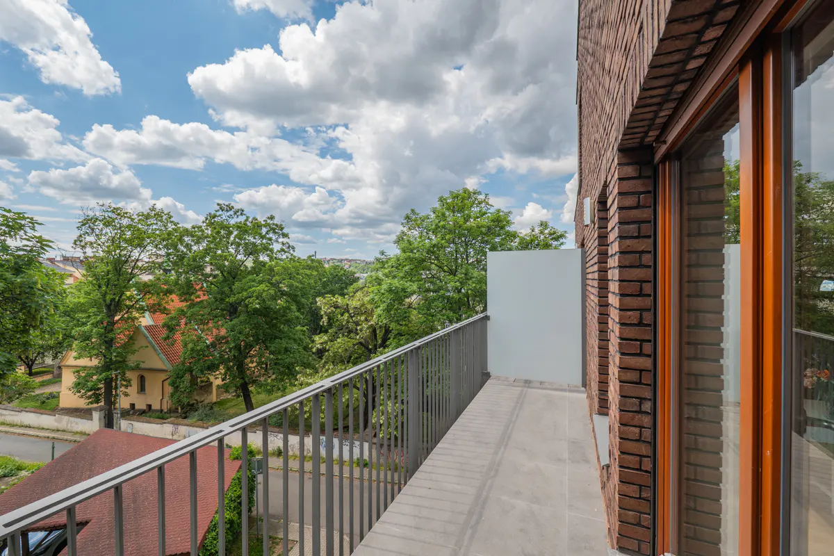 Balcony view of a brick building with metal railings overlooking green trees and a cloudy sky.