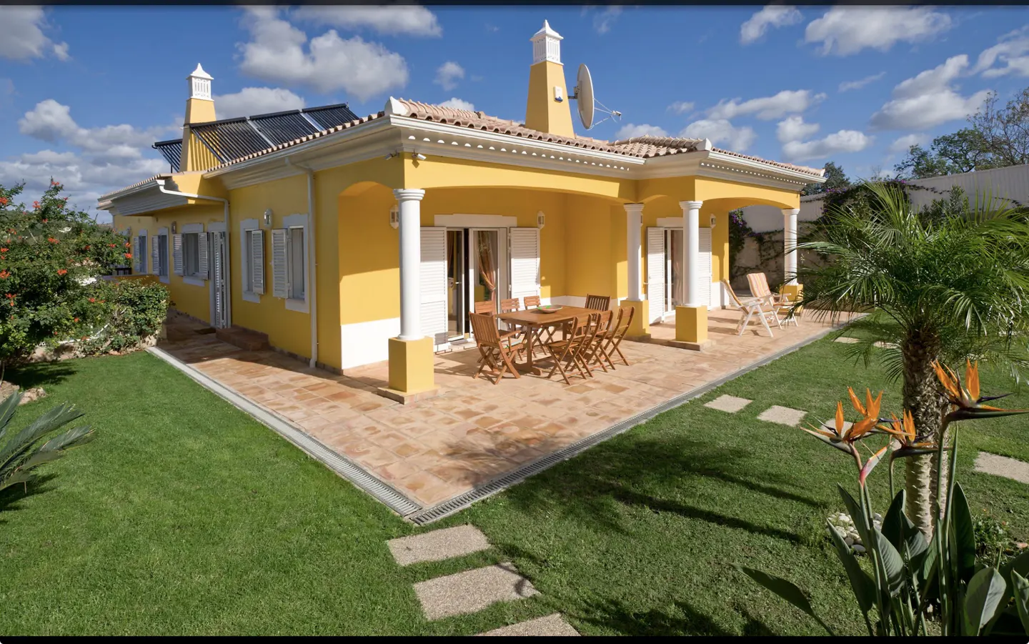 Bright yellow single-story villa with a tiled patio, outdoor dining set, and lush garden under a sunny blue sky.