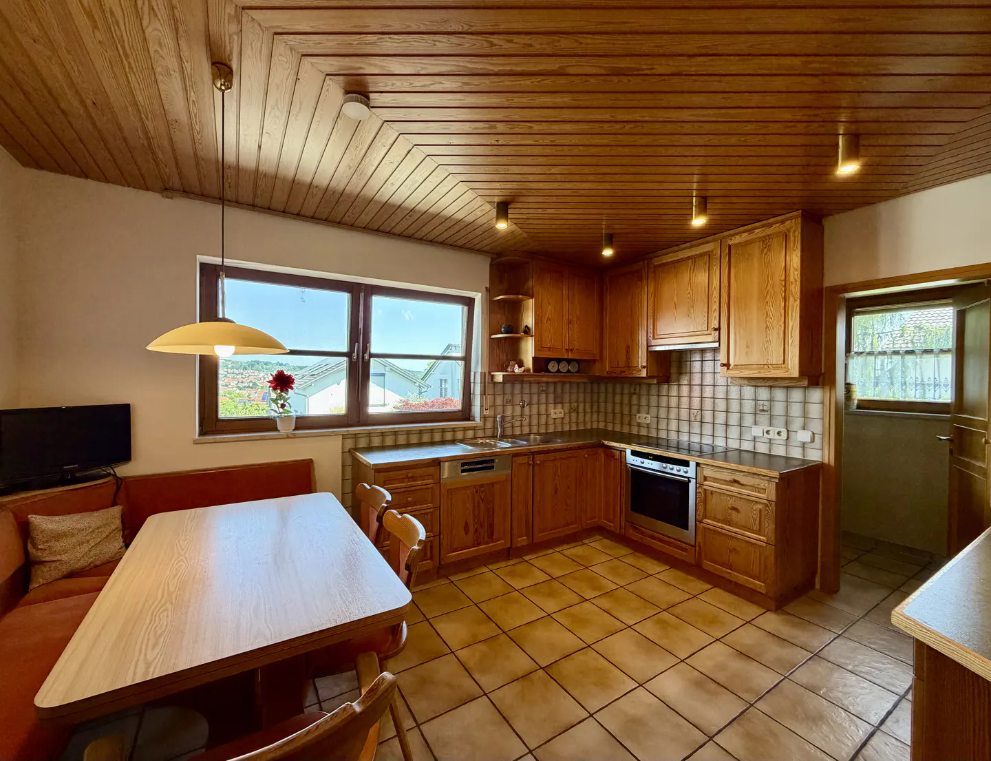 A kitchen with wood cabinets, a table with chairs, and a built-in bench. The ceiling is wood paneling. A window is visible.