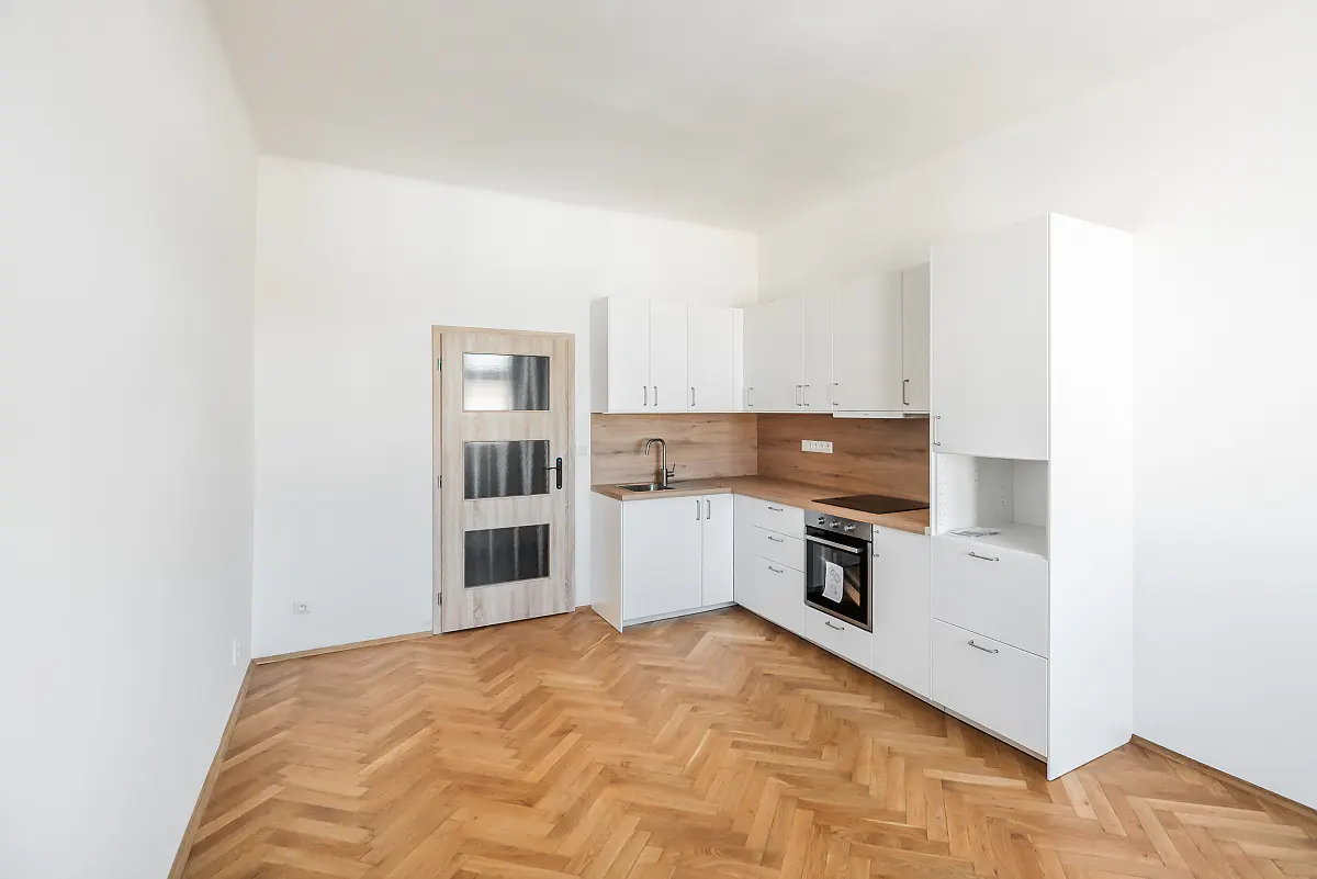 Bright, empty kitchen with white cabinets, wood countertops, and herringbone wood floors. A light wood door is on the left.