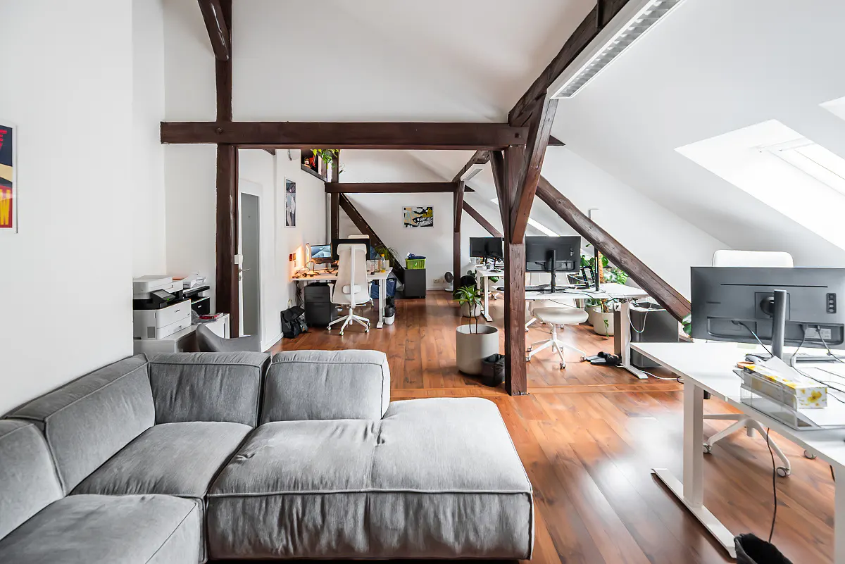 Open office space with exposed beams, hardwood floors, and white walls. A gray sectional sofa is in the foreground. Desks with computers are in the background.
