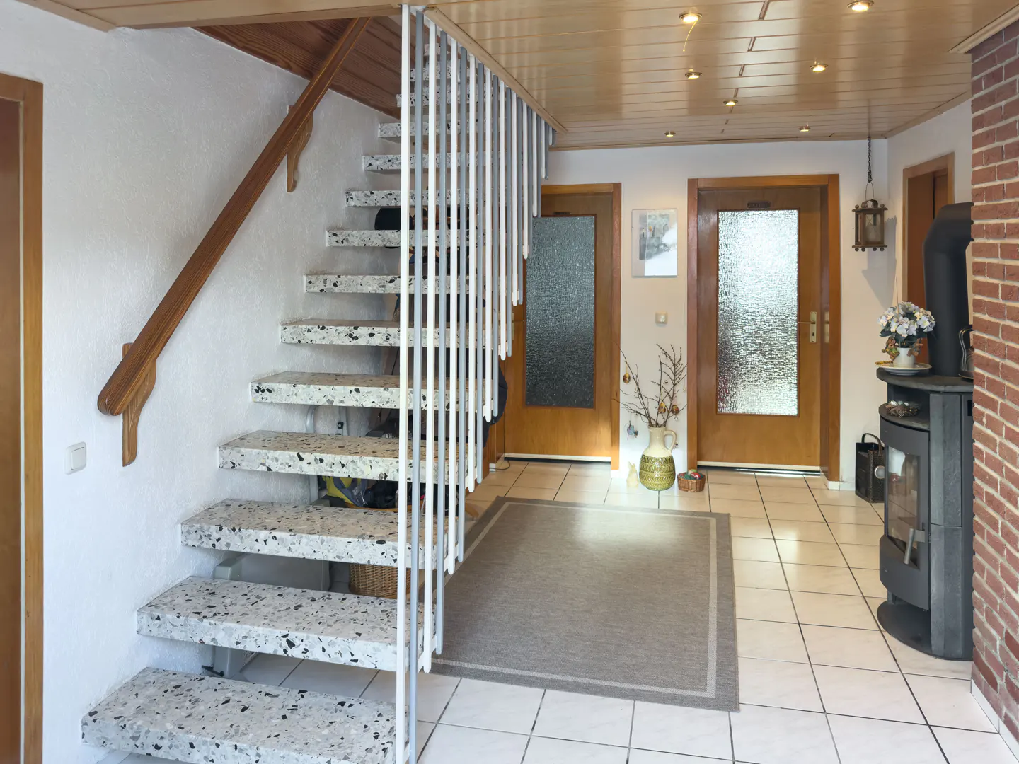 Entryway with terrazzo stairs, white railings, and a wood handrail. A gray rug sits on white tile floors. A wood stove is near a brick wall.