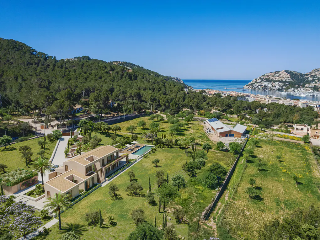 Aerial view of a modern beige house with a pool, surrounded by green lawns, trees, and a forest-covered hill, with a blue sky and ocean in the background.