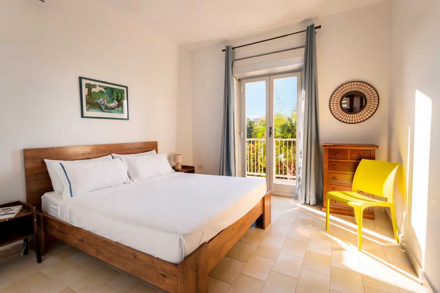 Bright bedroom with a wooden bed, white linens, and a balcony. A yellow chair sits near a wooden dresser and round mirror.