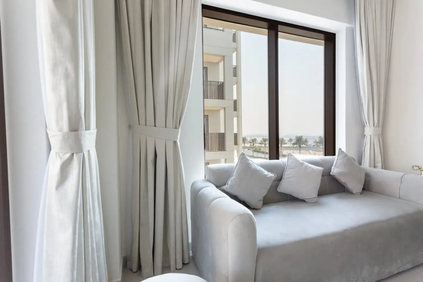 Bright living room with a gray sofa, white pillows, and curtains. A large window shows a building and palm trees in the distance.