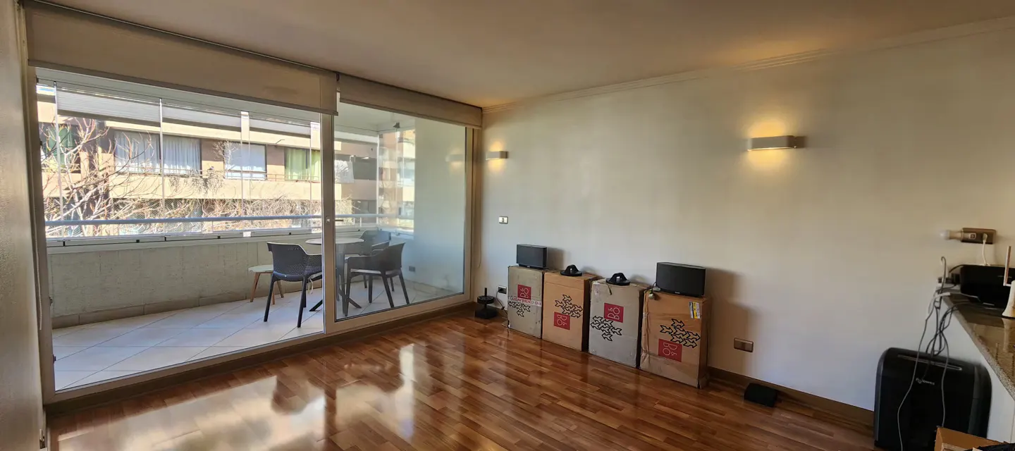 Bright living room with hardwood floors, a balcony with chairs and table, and boxes with speakers on top against the wall.