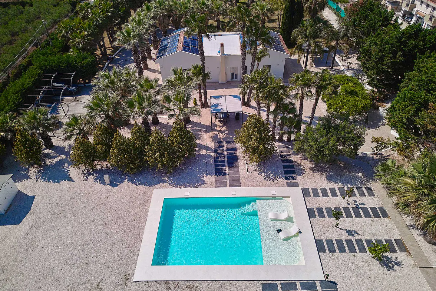 Aerial view of a white villa with solar panels, a turquoise pool with lounge chairs, and palm trees on a gravel lot.