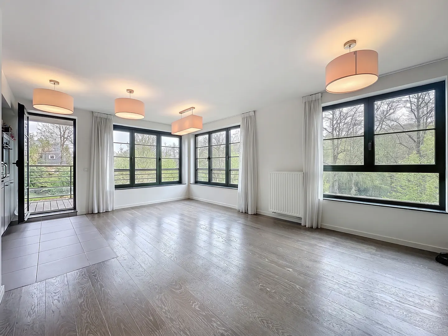 Bright, empty room with gray wood floors, white walls, and black-framed windows. Balcony door open to greenery. Four drum pendant lights illuminate the space.