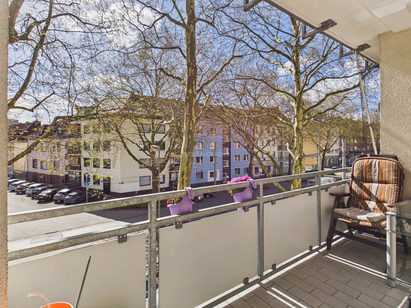 Balcony view with potted pink flowers on the railing, a chair, and a small table. Buildings and trees are visible in the background.