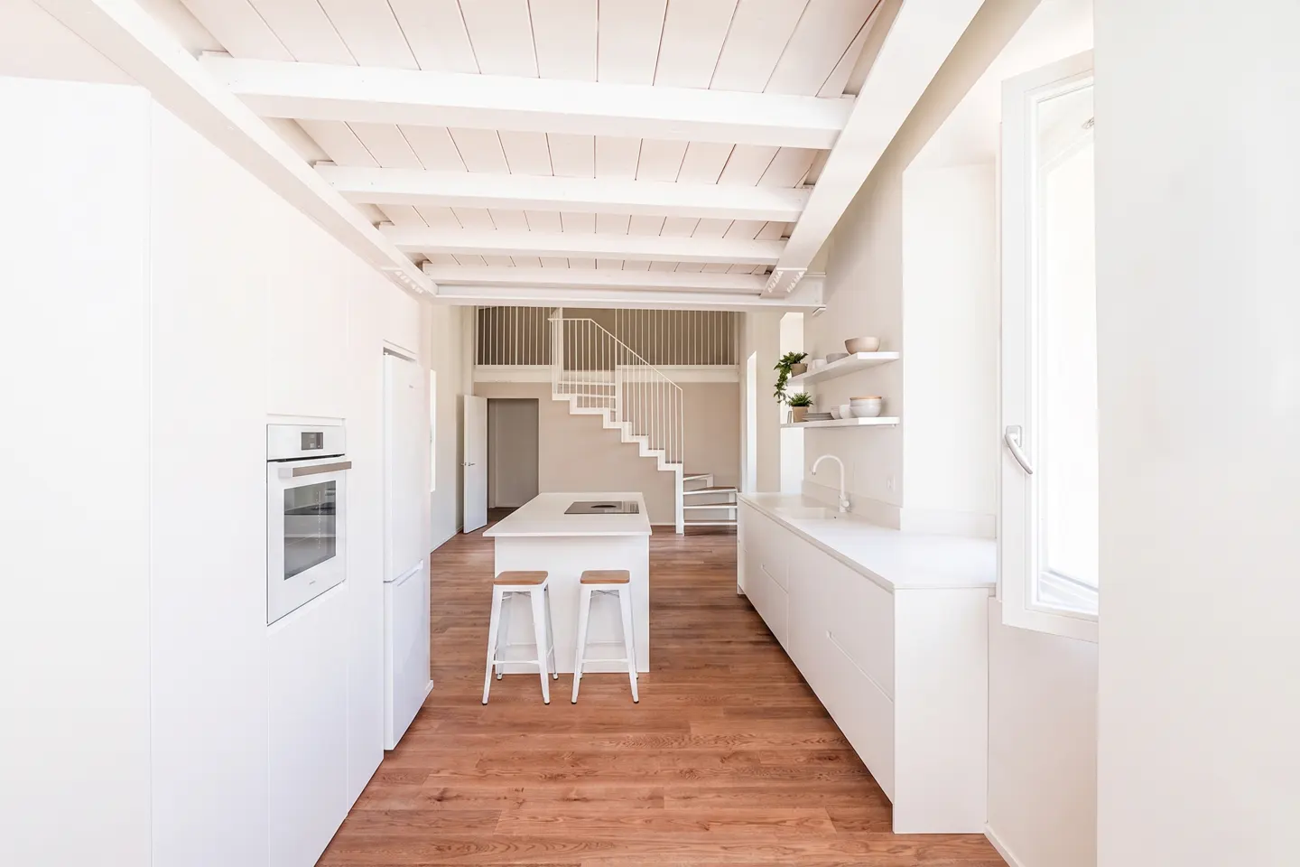 Bright, modern kitchen with white cabinets, island with stools, and wood floors. A white staircase leads to a loft area.