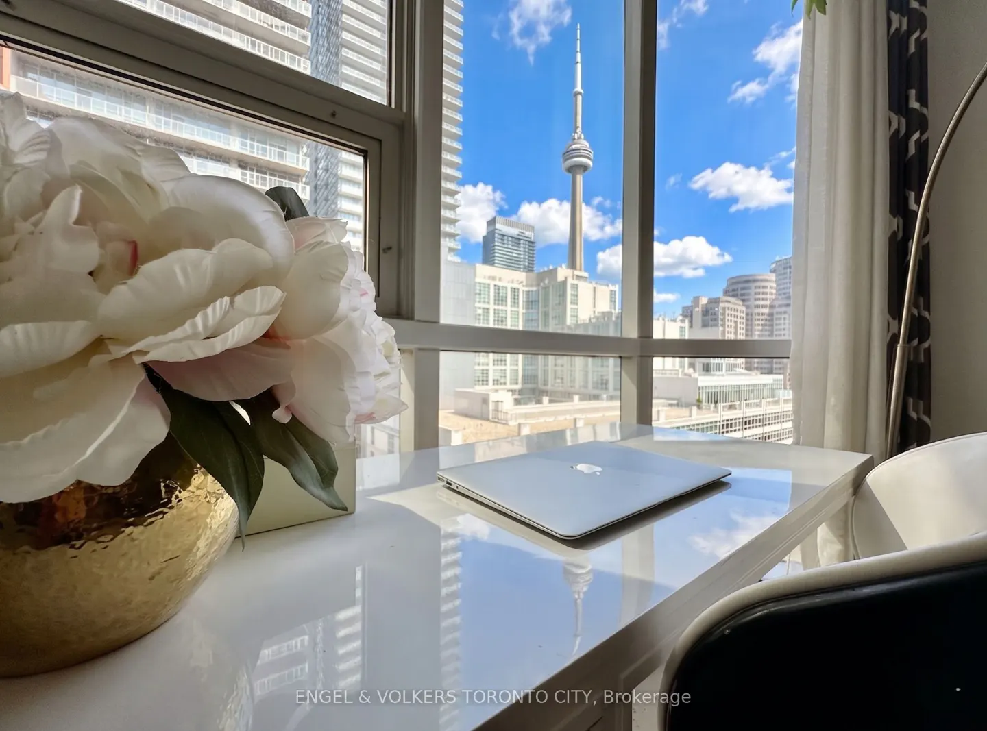 A white desk with a laptop and a gold vase of white flowers overlooks the Toronto skyline and CN Tower.