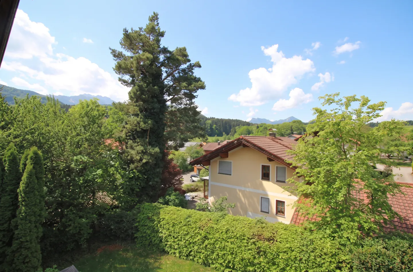View of a yellow house with a red tile roof, surrounded by green trees and a blue sky with white clouds. Mountains are visible in the distance.