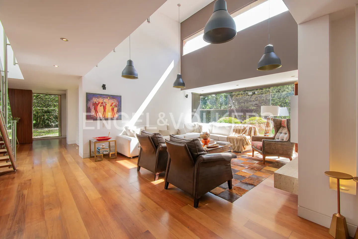 Modern living room with hardwood floors, leather armchairs, a white sofa, and large windows overlooking a garden.