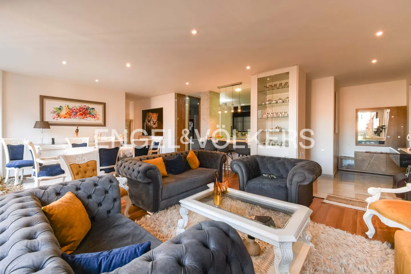 A wide shot of a living room with gray sofas, blue and yellow pillows, and a glass coffee table. A dining area is in the background.
