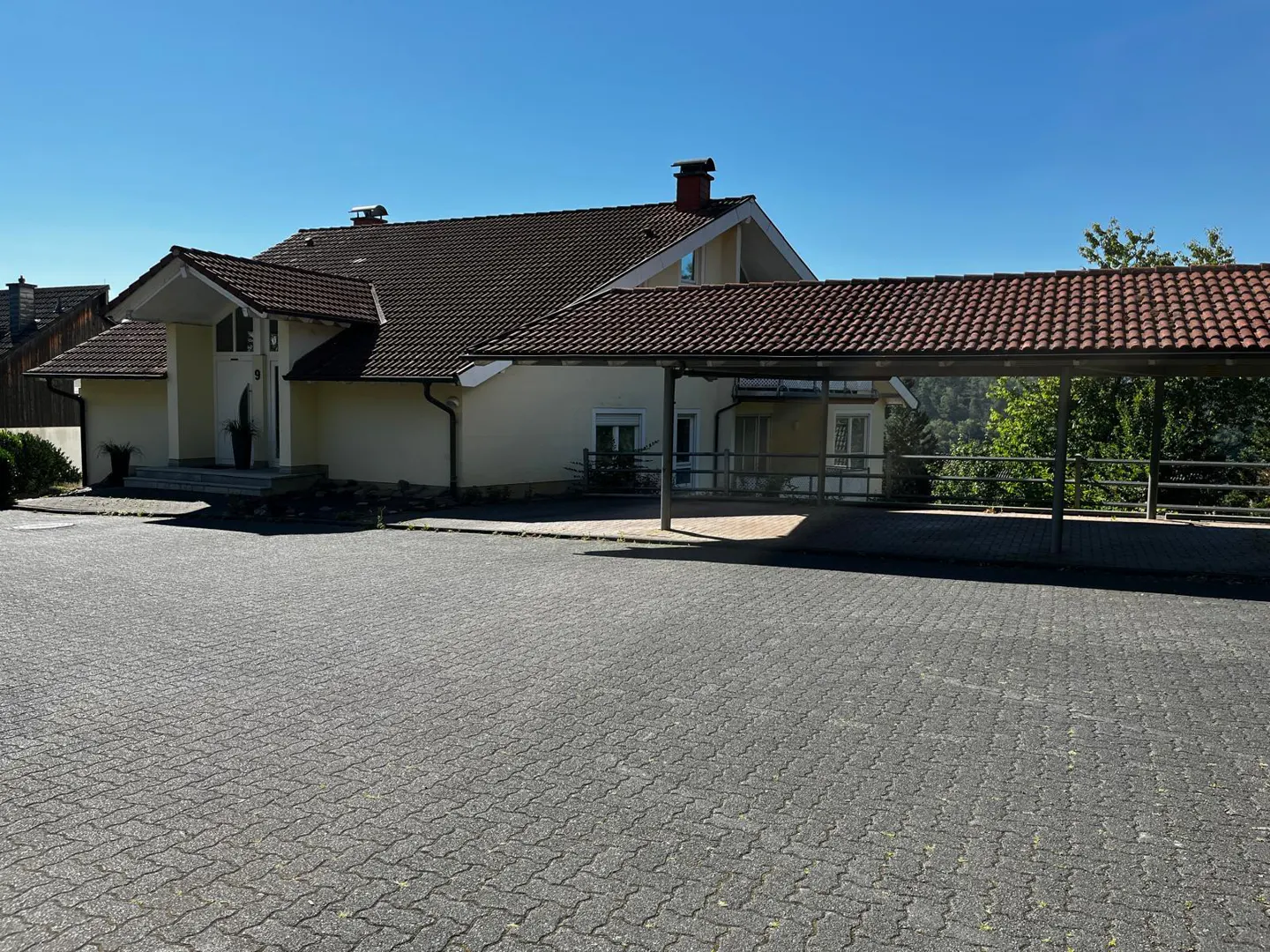 Beige house with a brown tile roof and a carport on a paved driveway under a clear blue sky.